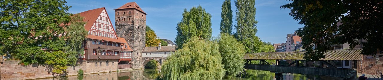 Blick auf mittelalterliche Gebäude Nürnbergs im Sonnenschein, links ein Fachwerkhaus, dann eine Steinturm, eine bebaute Steinbrücke über die Pegnitz, Bäume und ein überdachter Holzsteg