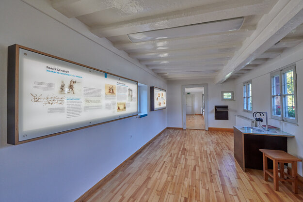 Blick in einen Ausstellungsraum im Henkerhaus, links eine Informationstafel an der Wand, rechts eine Hörstation mit Tisch, Stühlen und Kopfhörer