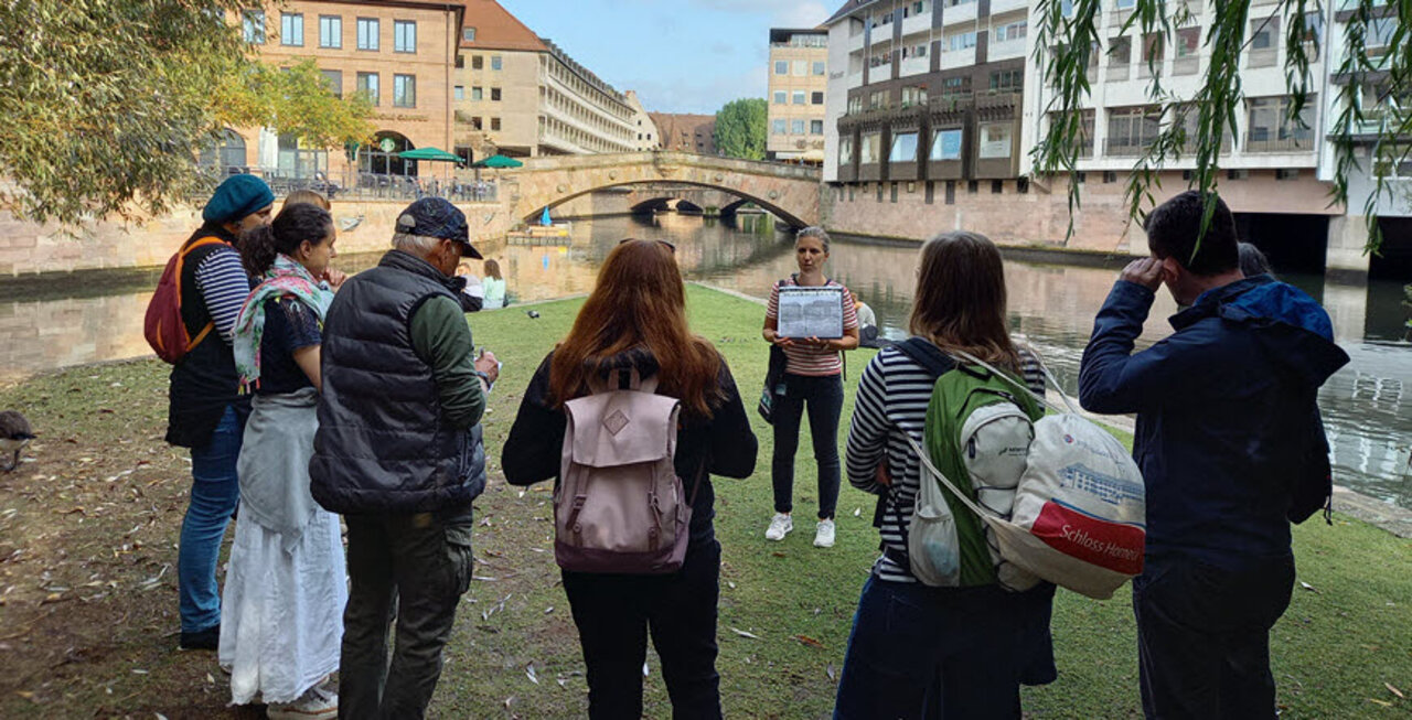 Szene bei einer Stadtführung, die Gäste sind von hinten zu sehen und die Stadtführerin hält einen Ordner mit Bildern hoch, im Hintergrund ein Fluss mit Steinbrücke
