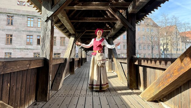 Frau in historischem Kostüm steht lächelnd auf einer Holzbrücke, die Arme einladend ausgebreitet